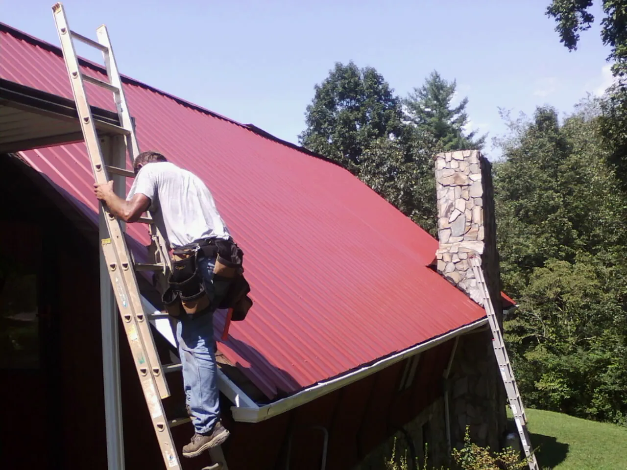 Roofer on a ladder beside a red metal roof and stone chimney — Soffit & Fascia Repair by Roofing Brothers in Glastonbury